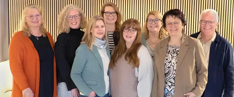 Personen gruppiert vor Lamellenwand, eine Person vorne sitzend, alle lächelnd in hellen und dunklen Outfits.